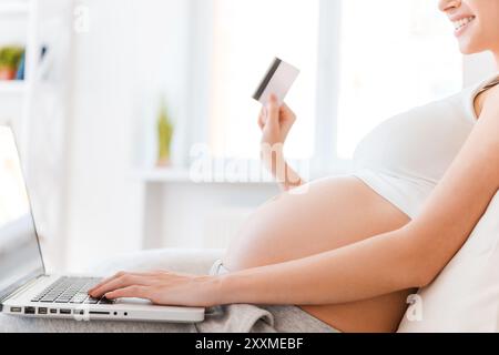 Shopping online. Vista laterale della giovane donna incinta che tiene in mano una carta di credito e un'altra sul laptop mentre è seduta sul divano Foto Stock