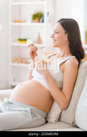 Mantenere la calma mentre si è incinta. Sorridente giovane donna incinta che mangia yogurt e guarda lontano mentre si siede sul divano Foto Stock