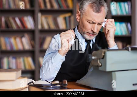 E' tutto sbagliato! Frustrato uomo anziano di capelli grigi in abbigliamento formale seduto alla macchina da scrivere e tenendo in mano pezzi di carta con la libreria Foto Stock