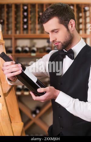 Sommelier che sceglie il vino. Giovane sicuro di sé con gilet e cravatta che tiene la bottiglia con il vino e la guarda mentre si trova in un negozio di liquori Foto Stock