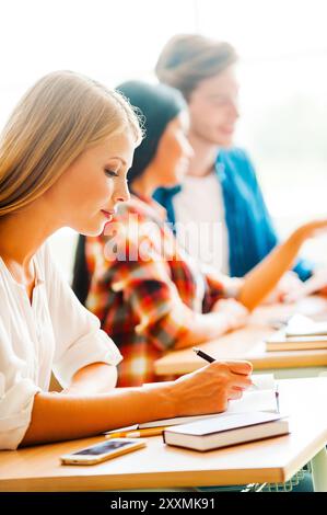 Lavorare sodo per un buon futuro. Giovane donna concentrata che scrive in un blocco appunti mentre si siede alla sua scrivania in classe Foto Stock