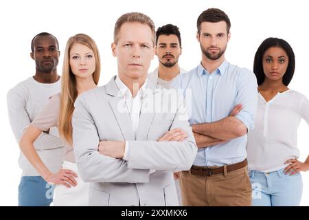 Guidare la sua squadra verso il successo. Uomo maturo e sicuro in un abbigliamento casual elegante, che tiene le braccia incrociate e guarda la fotocamera mentre un gruppo di giovani è in piedi Foto Stock