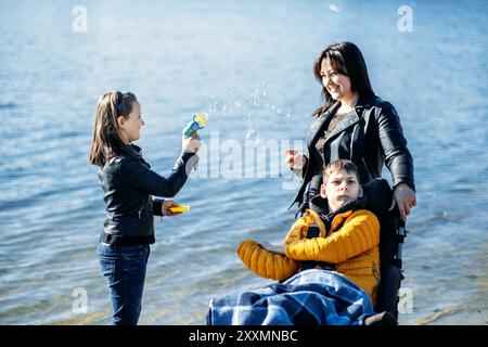 Mamma e bambini si divertono all'aperto vicino al lago, attività per famiglie incluse, bambino in sedia a rotelle, bolle d'aria dei fratelli, inclusione disabili Foto Stock