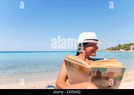 Ritratto di una donna felice seduta su una sdraio e a leggere un libro. Donna felice sulla spiaggia con una vista bellissima. Concetto di vacanza estiva. Foto Stock