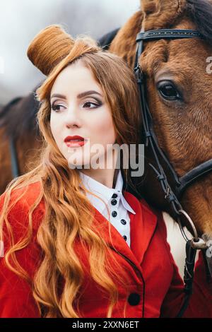 ragazza di fantino dai capelli rossi in un cardigan rosso e stivali neri alti con un cavallo per una passeggiata Foto Stock