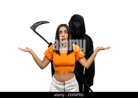 Grim reaper, vestito con un costume di Halloween con un mantello scuro e un volto scheletrico, tiene una falce mentre si intrufola dietro una giovane donna bruna terrorizzata Foto Stock