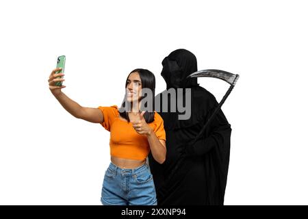 Grim reaper, vestito con un costume di Halloween con un mantello scuro e un volto scheletrico, tiene una falce in una mano mentre scatta un selfie con uno smartphone al Foto Stock