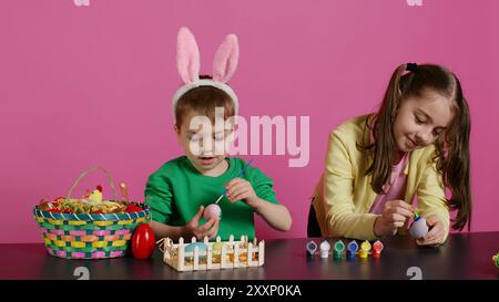 Bambini piccoli felici che usano acquerelli e francobolli per dipingere le uova in tempo per le festività di pasqua, sfondo rosa. Allegri piccoli fratelli che decorano gli ornamenti con disegni creativi. Telecamera B.. Foto Stock