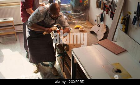 Scultore che forma il legno grezzo con scalpello e martello in falegnameria con banco di lavoro per la pulizia dei collaboratori in background. Uomo BIPOC che produce sculture in legno, tavola per incisione con utensili, telecamera B. Foto Stock