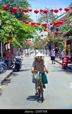 Hoi An, Vietnam - 15 aprile 2009: Una donna in bicicletta percorre la colorata strada principale di Hoi An, in Vietnam al sole di mezzogiorno. Foto Stock