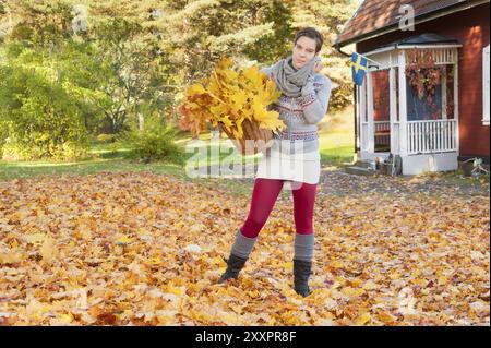 Attraente giovane donna con cesto pieno di foglie autunnali di fronte a una tipica casa svedese in legno dipinta di rosso. Donna attraente che colleziona l'autunno Foto Stock
