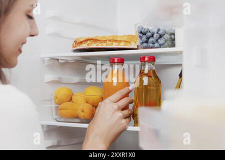 Donna che prende, afferra o raccoglie la bottiglia di vetro del succo dalla mensola aperta del frigorifero o dal cassetto del frigorifero pieno di frutta, verdura, banana, pesche Foto Stock