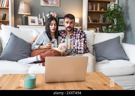 Una coppia seria guarda la TV insieme mentre si siede su un divano nel soggiorno. Fidanzata e fidanzato abbracciano, coccolano, parlano e guardano la TV St Foto Stock