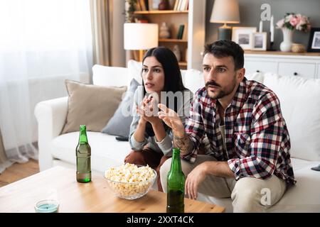 Una coppia seria guarda la TV insieme mentre si siede su un divano nel soggiorno. Fidanzata e fidanzato abbracciano, coccolano, parlano e guardano la TV St Foto Stock