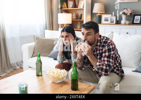 Una coppia seria guarda la TV insieme mentre si siede su un divano nel soggiorno. Fidanzata e fidanzato abbracciano, coccolano, parlano e guardano la TV St Foto Stock