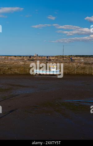 EDIMBURGO - 28 LUGLIO 2024: Una barca da pesca è ancorata con la bassa marea mentre i ciclisti viaggiano lungo le mura del porto di Fisherrow sotto un cielo estivo azzurro. Foto Stock