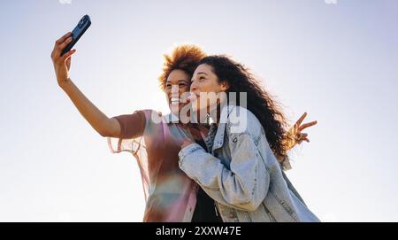 Due giovani migliori amici si mettono in posa e scattano un selfie all'aperto in una giornata di sole, catturando insieme i loro momenti felici e felici. Foto Stock