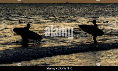 Salvador, Bahia, Brasile - 6 dicembre 2019: Due surfisti sono visti sul bordo della spiaggia di Farol da barra durante il tramonto. Salvador, Bahia. Foto Stock