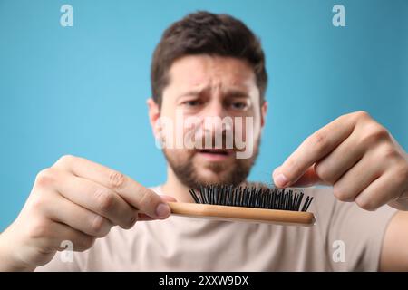 Uomo triste che prende i capelli persi dalla spazzola su sfondo azzurro, messa a fuoco selettiva. Problema di alopecia Foto Stock