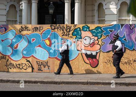 Notting Hill, Londra, Regno Unito. 26 agosto 2024. Il più grande festival di strada d'Europa si svolge nelle strade di Notting Hill. Ballerini esotici a tema giamaicano e gruppi musicali sfilano per le strade, con cibo e intrattenimento di strada intorno alla zona che si aggiungono all'evento. La Grand Parade si svolge il lunedì festivo come il culmine del festival di tre giorni, iniziato nel 1966. Agenti di polizia che passano graffiti da clown sugli accumuli di sicurezza che proteggono le proprietà Foto Stock