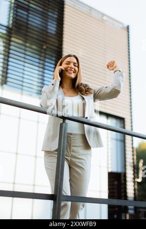 Una donna d'affari gioiosa festeggia un momento all'aperto, partecipando a una allegra conversazione telefonica mentre si appoggia a una ringhiera accanto a un elegante buildi Foto Stock