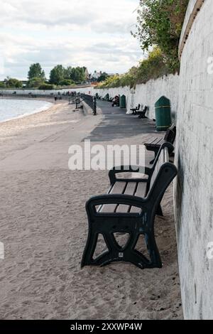 BURNTISLAND, FIFE, SCOZIA, Regno Unito - 11 GIUGNO 2024: Le panche sono allineate lungo una spiaggia sabbiosa vicino all'acqua, con persone sedute in lontananza. Foto Stock