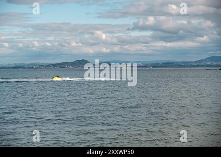 BURNTISLAND, FIFE, SCOZIA, Regno Unito - 11 GIUGNO 2024: La vivace moto d'acqua gialla si muove sull'acqua serena, mostrando lo skyline della città di Edimburgo sullo sfondo. Foto Stock