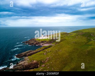 Una prospettiva aerea cattura il faro di Rubha Réidh alto tra scogliere spettacolari e verde ondulato, affacciato sul vasto mare blu. Foto Stock