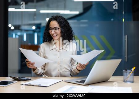 Una donna d'affari si siede alla scrivania dell'ufficio per esaminare i documenti. Un professionista sorridente si occupa di multitasking con notebook e telefono, per una produttività ottimale nello spazio di lavoro creativo. Foto Stock