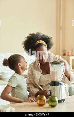 Donna afroamericana sorridente riccia che prepara frullati di latte usando il frullatore, mentre la bambina allegra sussurra qualcosa nell'orecchio delle mamme nella cucina beige, copia spazio Foto Stock