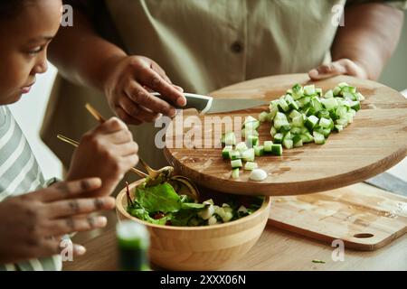 Primo piano sulle mani di una donna anziana nera che aggiunge cetrioli tritati alle foglie di lattuga verde nel recipiente dell'insalata utilizzando un tagliere di legno e un coltello mentre cucinano insieme alla ragazza in cucina, copiare lo spazio Foto Stock