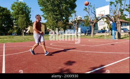 Un anziano vecchio italiano gioca a basket su un campo sportivo in una giornata estiva. Foto Stock