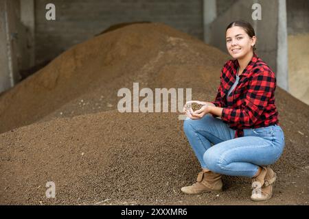 Giovane lavoratrice dell'azienda lattiero-casearia che accovacciava un mucchio di buccia di soia nel deposito dei foraggi Foto Stock