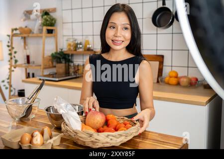 Una attraente blogger asiatica di cibo sta cucinando davanti alla macchina fotografica, registrando un video di se stessa che prepara cibo sano nella sua cucina di casa. Foto Stock