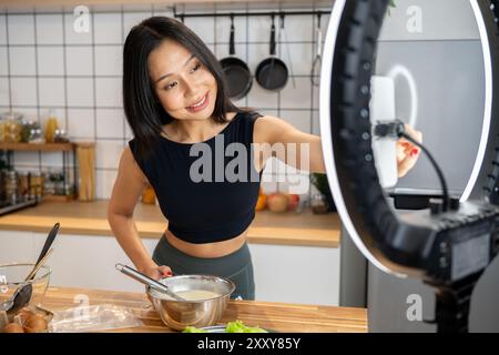 Una bella blogger di cibo asiatica o influencer sta registrando se stessa o andando in diretta sui social media mentre prepara un piatto sano in cucina, adj Foto Stock