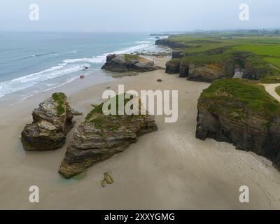 Ampia spiaggia con grandi formazioni rocciose sparse e acque calme, delimitata da ripide scogliere costiere, vista aerea, Praia das Catedrais, Playa de las Cated Foto Stock