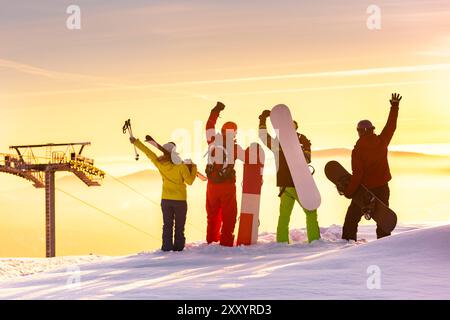 Diversi gruppi di giovani amici felici sciatori e snowboarder sono in piedi sulla cima della montagna al tramonto vicino agli impianti di risalita e godono di una splendida vista. Concetto di stazione sciistica Foto Stock