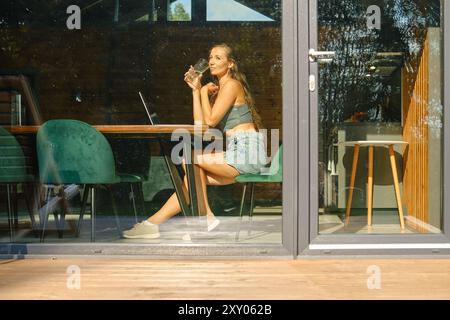 Vista dalla finestra di una donna che guarda un video su un laptop e sorseggia acqua minerale Foto Stock