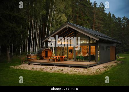 Una cabina moderna con grandi finestre in vetro e terrazza in legno con mobili da patio in una radura forestale e una sauna in legno a forma di botte Foto Stock