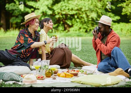 Un gruppo di amici seduto sulla coperta nel parco verde per fare un picnic con un assortimento di cibo, mentre uno scatta foto con la macchina fotografica, creando momenti indimenticabili Foto Stock