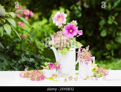 Bellissimo bouquet di fiori rosa, magenta hollyhock e ortensie viola in una teiera bianca in porcellana vintage sul tavolo. (Alcea rosea) Cottage gart Foto Stock