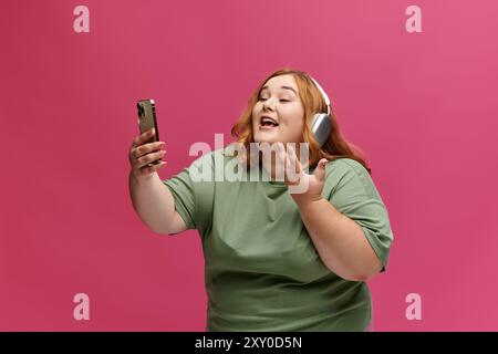 Una donna con i capelli rossi che indossa le cuffie canta entusiasticamente mentre tiene in mano uno smartphone. Foto Stock