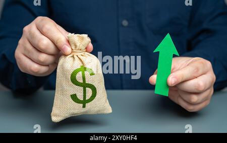Uomo che tiene in mano un sacco di soldi da dollari e una freccia verde che punta in alto, concetto. Aumento dei profitti e delle entrate. Stipendi e prestazioni. Budget più elevato, crescita delle pinne Foto Stock