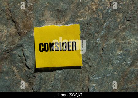 Considerare il messaggio scritto su un pezzo di carta strappato giallo con sfondo in pietra. Simbolo concettuale da considerare. Copia spazio. Foto Stock