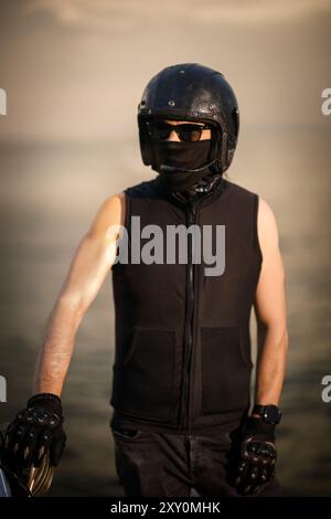 Ritratto di un motociclista che indossa un casco in pelle, occhiali di protezione e un rivestimento facciale, pronto per il viaggio. Il pilota è vestito di nero, dando un duro e myst Foto Stock