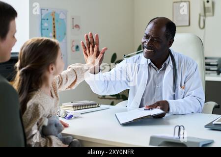 Medico afroamericano che vive una giovane ragazza durante una visita di routine con un genitore che osserva. Apparecchiature mediche e grafici in background, creando un'atmosfera coinvolgente e positiva in clinica Foto Stock