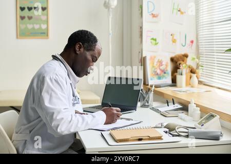 Medico afroamericano che scrive appunti alla scrivania in ufficio con attrezzature mediche e laptop visibile. L'ufficio è decorato con poster didattici e disegni per bambini che aggiungono calore Foto Stock