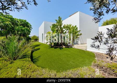 Questa immagine mostra una lussuosa villa bianca moderna circondata da un giardino tropicale splendidamente curato caratterizzato da palme, piante di banane e un Foto Stock