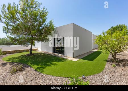 Una casa bianca contemporanea e minimalista caratterizzata da prato verde sintetico e alberi di pino e ulivo maturi, adagiata su un cielo blu limpido Foto Stock