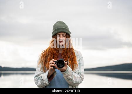 Una giovane donna con i capelli rossi esamina la sua macchina fotografica mentre si trova accanto a un lago sereno, indossando un berretto verde e una camicia a quadri, cattura la tranquillità Foto Stock